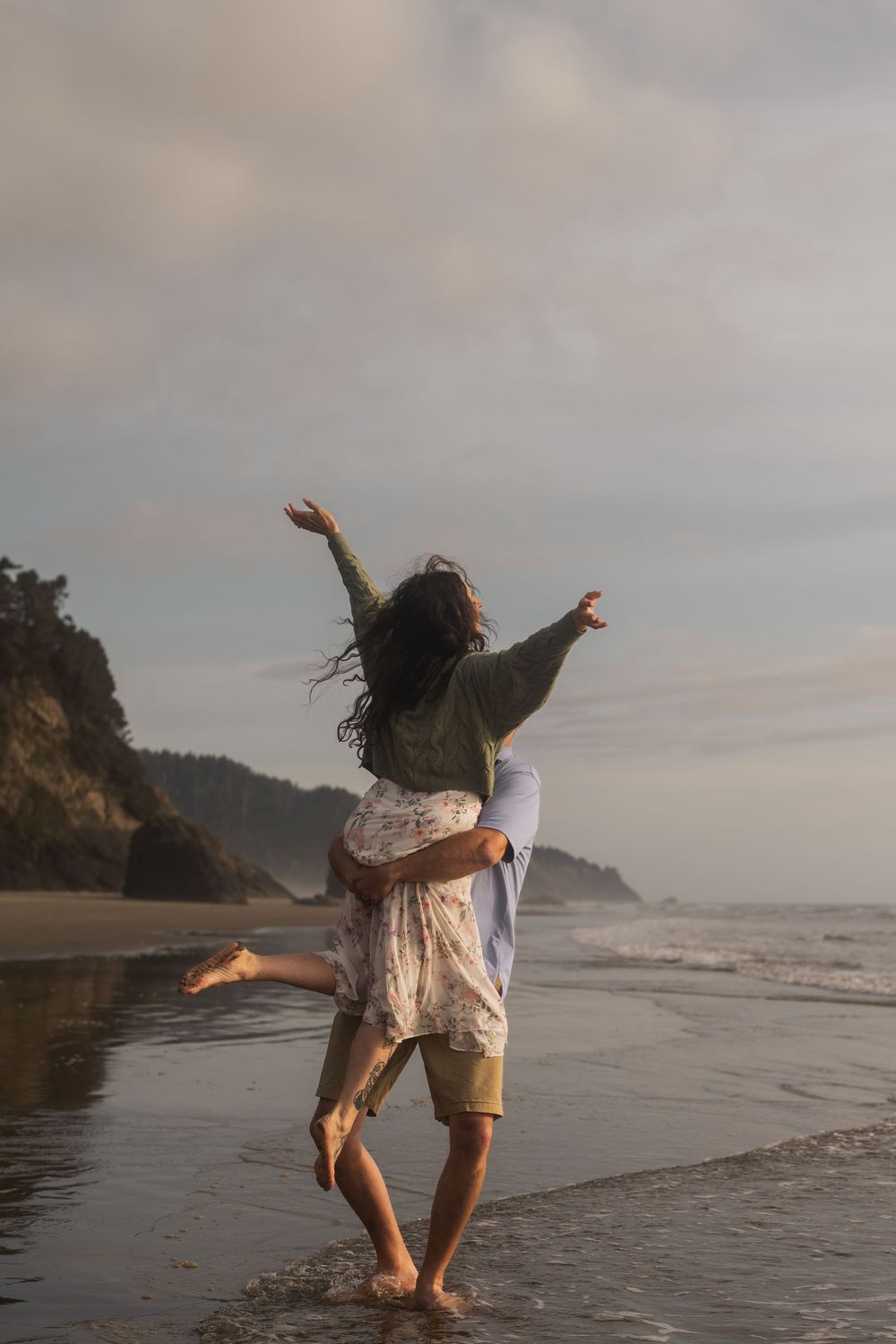A couple playfully dances on the beach with rugged coastal cliffs and moody sky in the background.