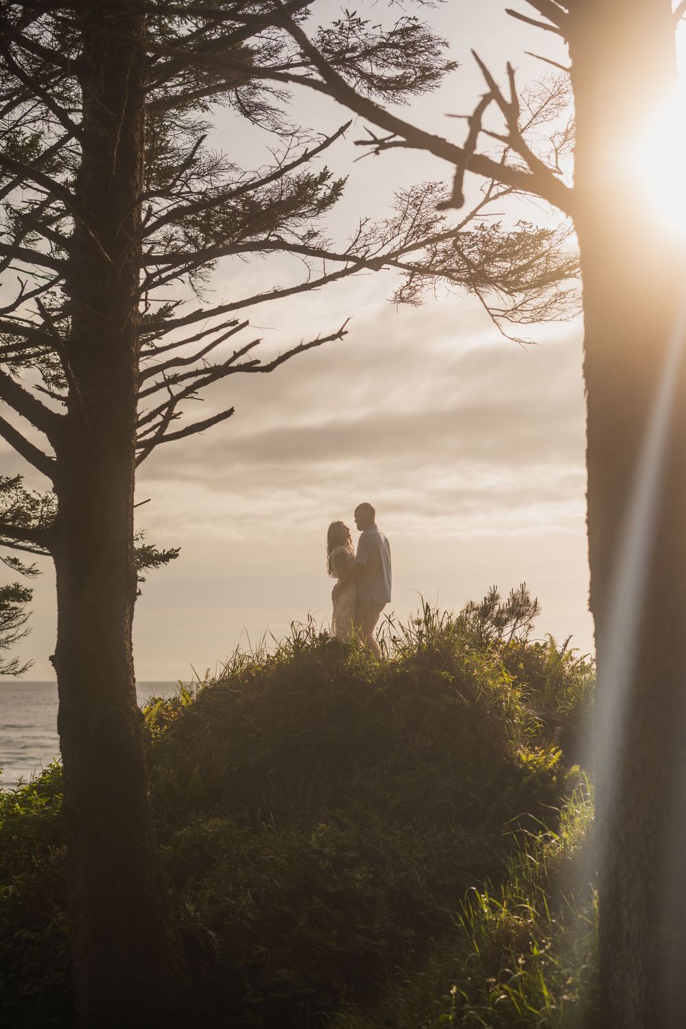 Silhouettes stand in embrace on a hilltop at sunset framed by dramatic tree branches.