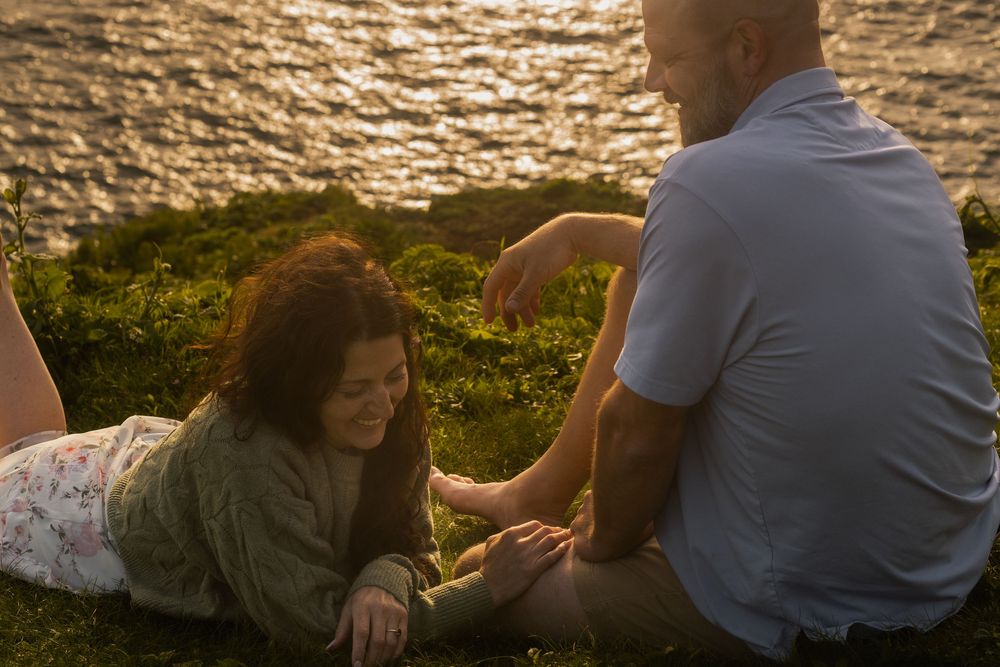 Two people share an intimate moment on a grassy cliff overlooking golden waters at sunset.