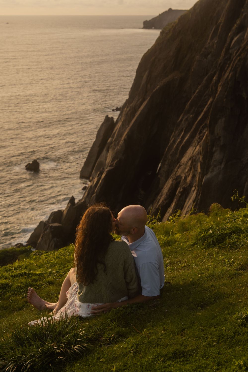 A couple sits together on a grassy cliff overlooking dramatic coastal rock formations during golden hour.