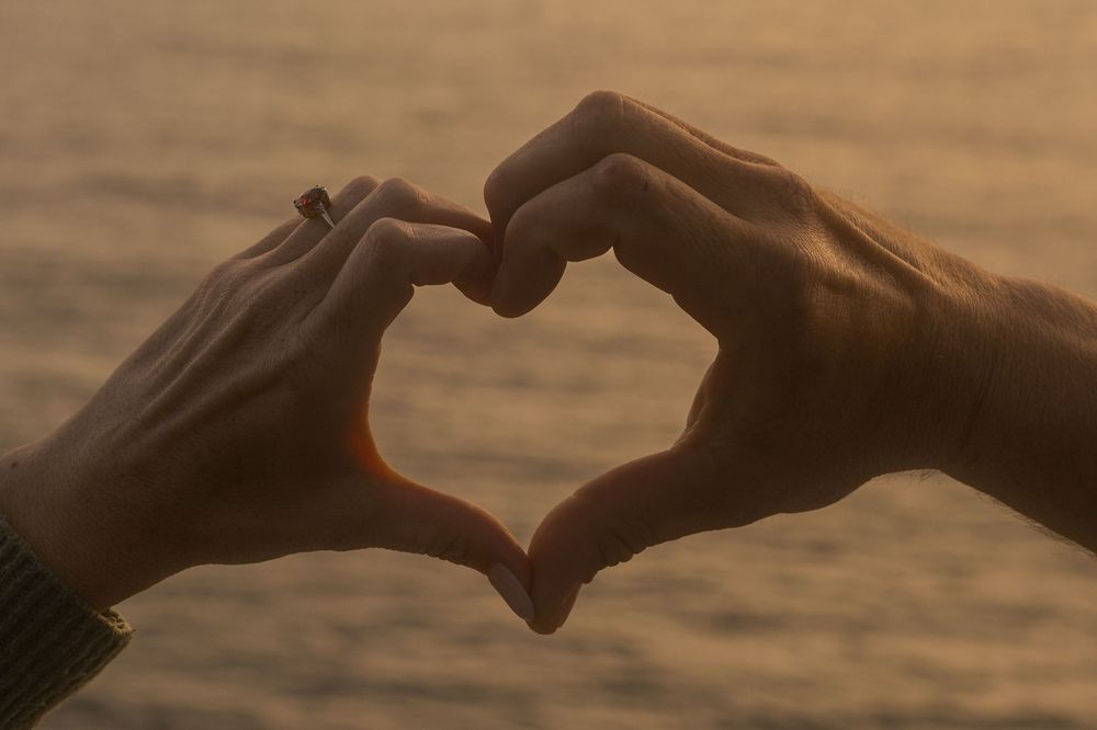 Two silhouetted hands forming a heart shape against a golden sunset background.