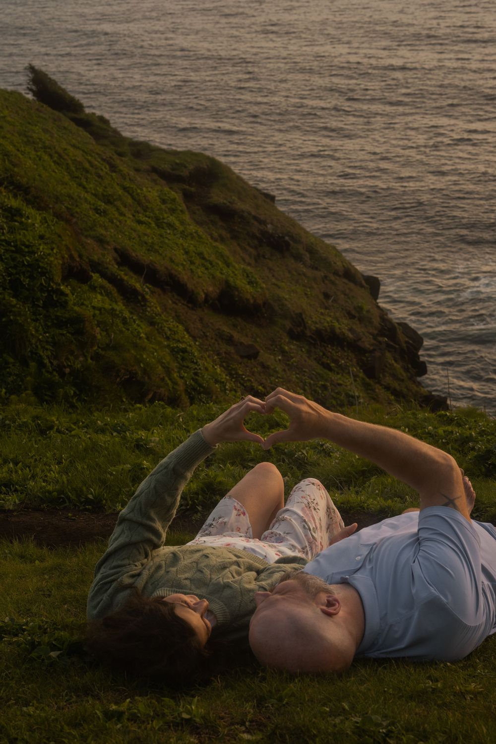 Two people lie on coastal grass overlooking the ocean at golden hour.