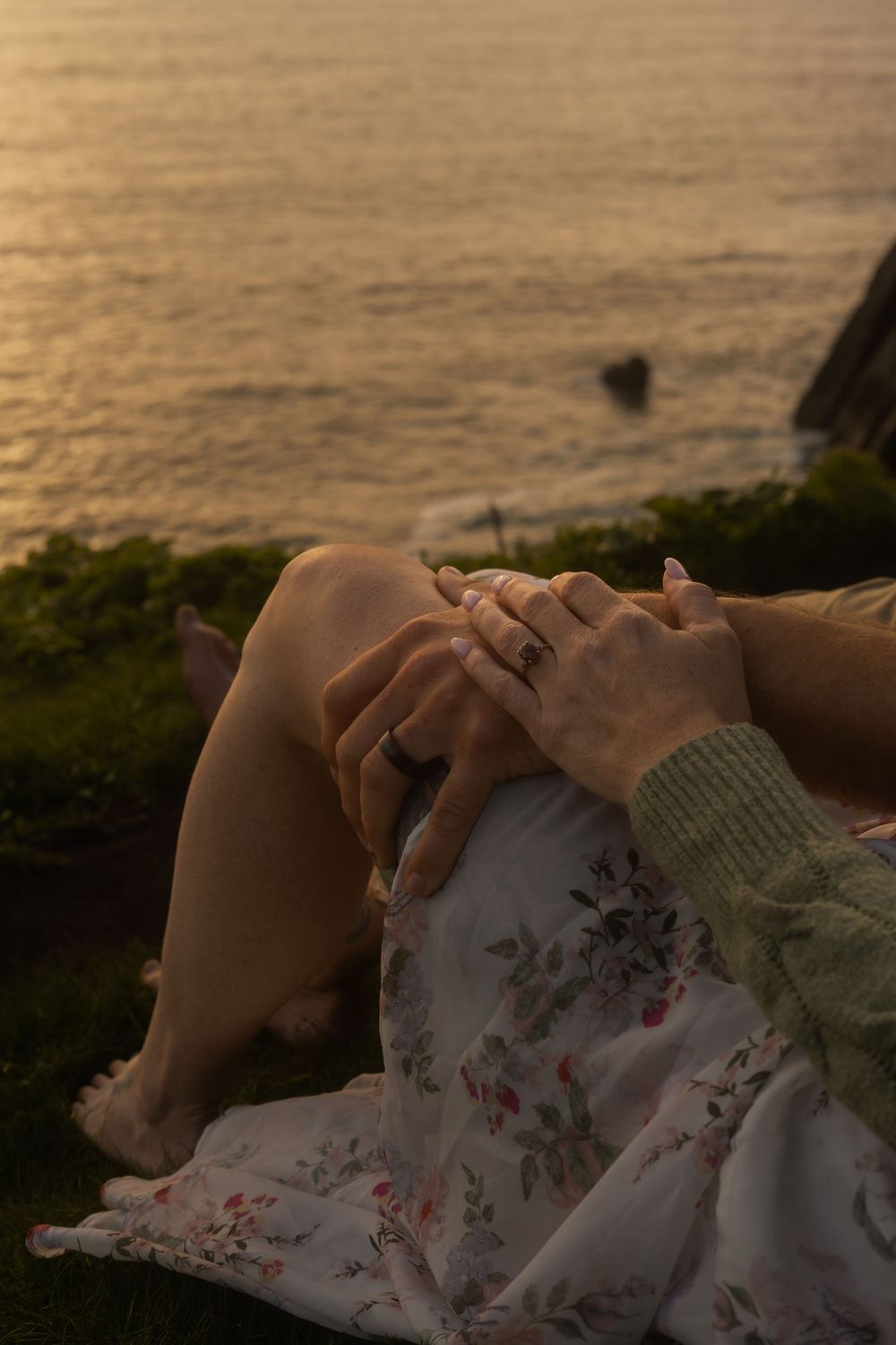 Close-up photo of hands clasped together while resting on a floral dress during sunset by the water.