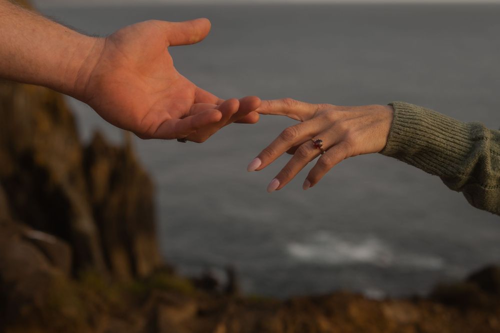 Two hands reach toward each other against a blurred ocean backdrop at sunset.