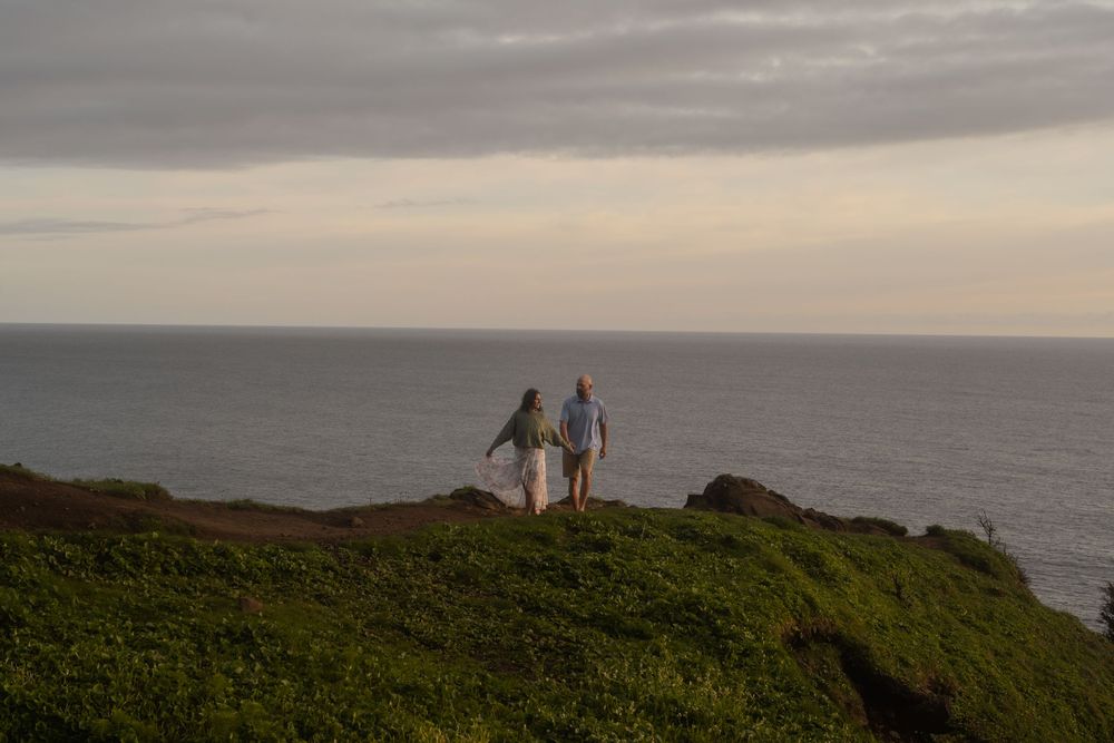Two figures stand together on a scenic coastal bluff overlooking the vast ocean at dusk.
