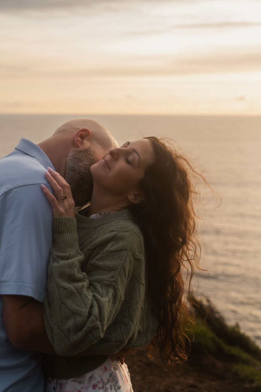 A couple embraces on a coastal cliff during golden hour with the ocean behind them.