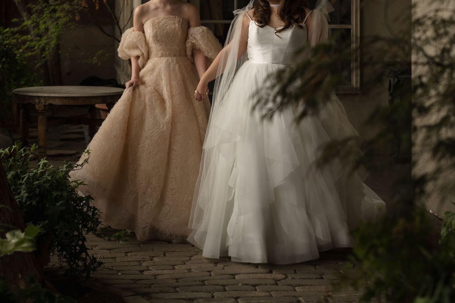 Two elegant ball gowns in champagne and white tulle photographed in a romantic garden setting with dappled sunlight.