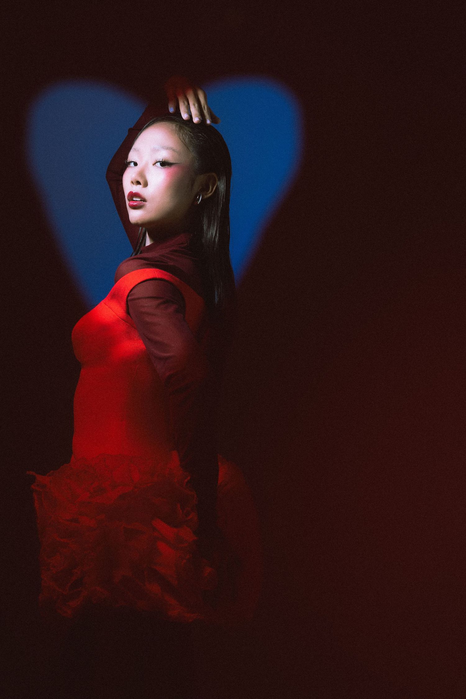 A figure in a red dress poses dramatically against a dark background with a large blue heart projection.