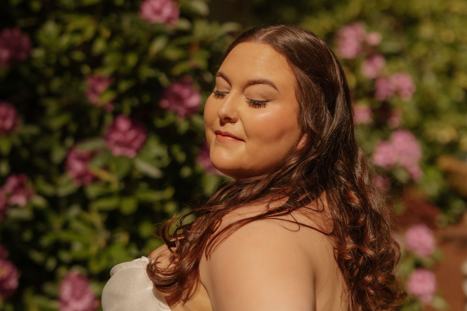 A person with long brown hair poses in sunlight with pink roses in the garden background.
