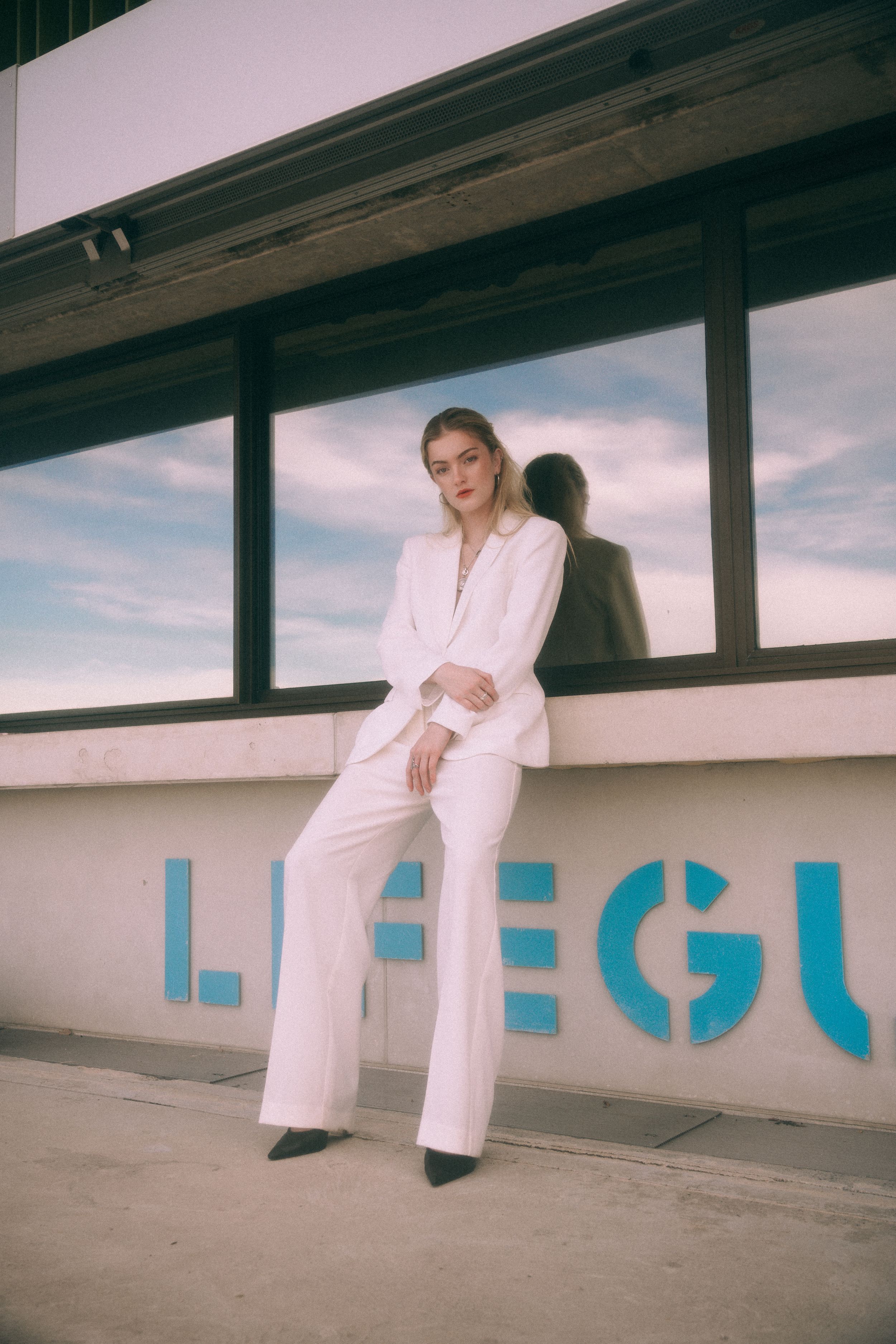 Person in a stylish white pantsuit poses against a wall with blue lettering at sunset.