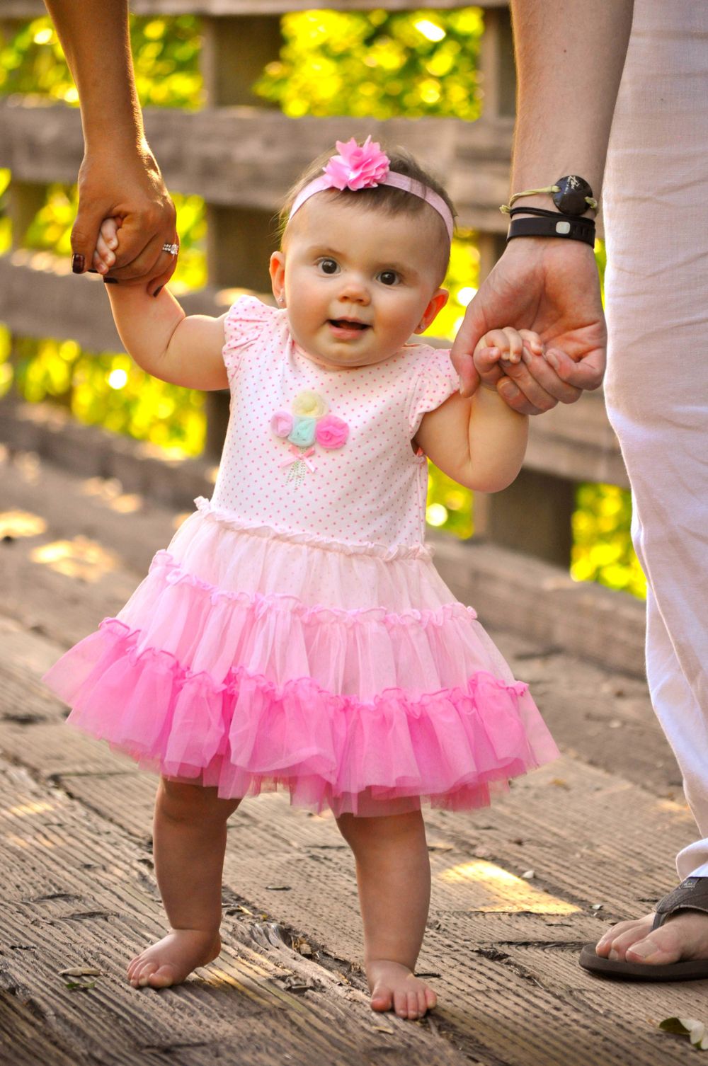 Baby in pink tulle dress taking first steps while holding parents' hands on wooden deck.