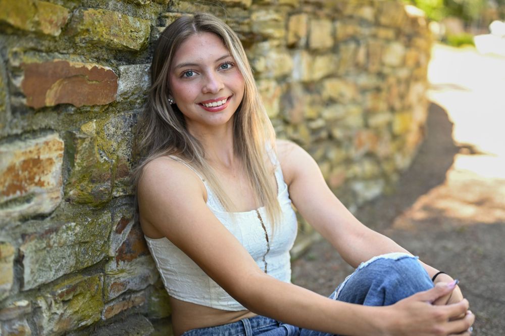 Casual portrait of someone sitting against a rustic stone wall in casual summer attire.