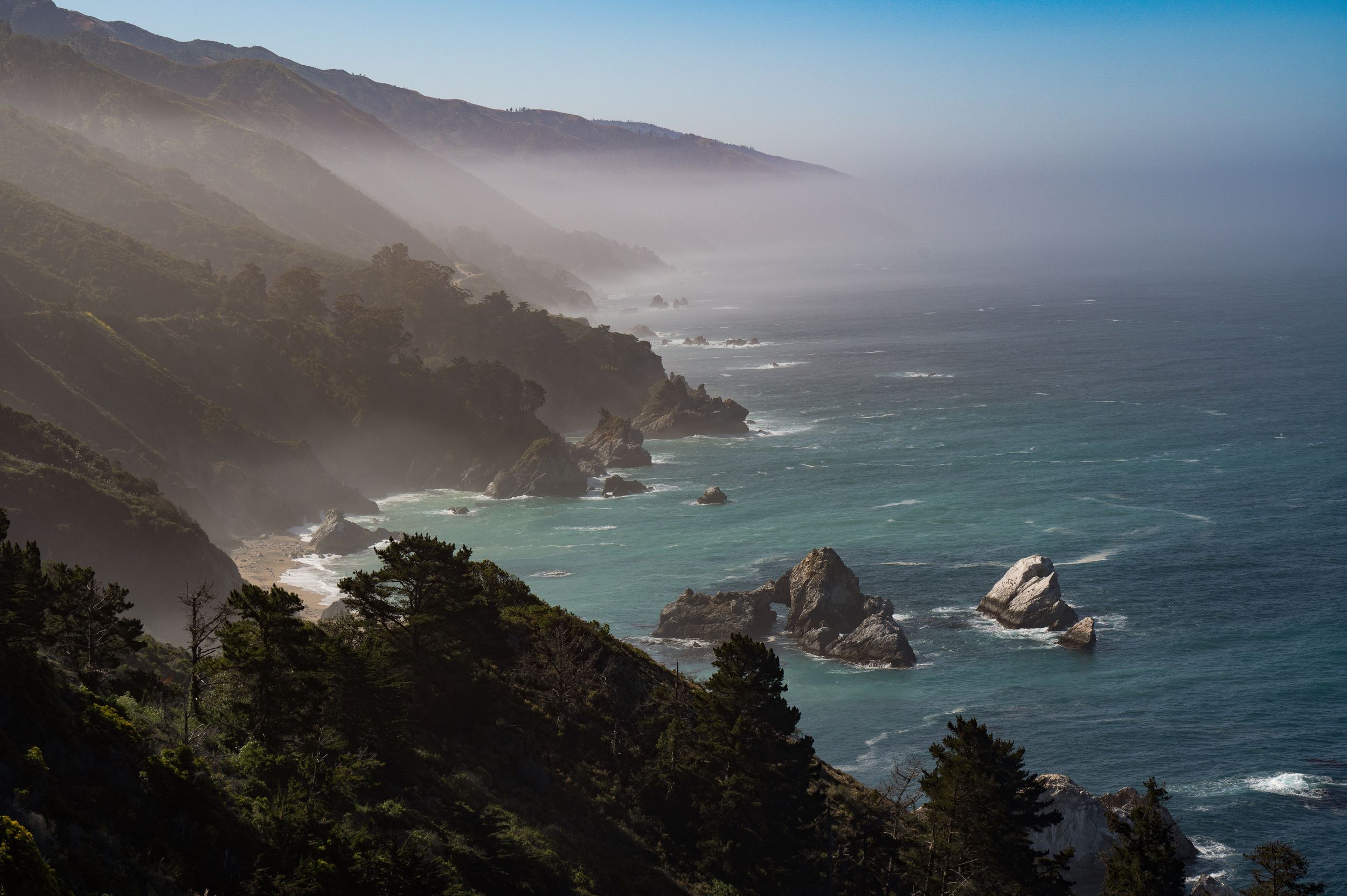 Foggy coastal landscape of Big Sur with rocky cliffs and ocean waves under misty conditions.