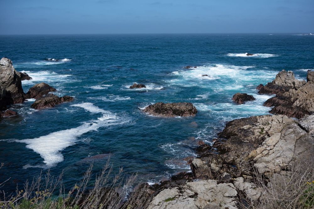 Rocky coastline with crashing waves against jagged rocks in deep blue ocean waters.