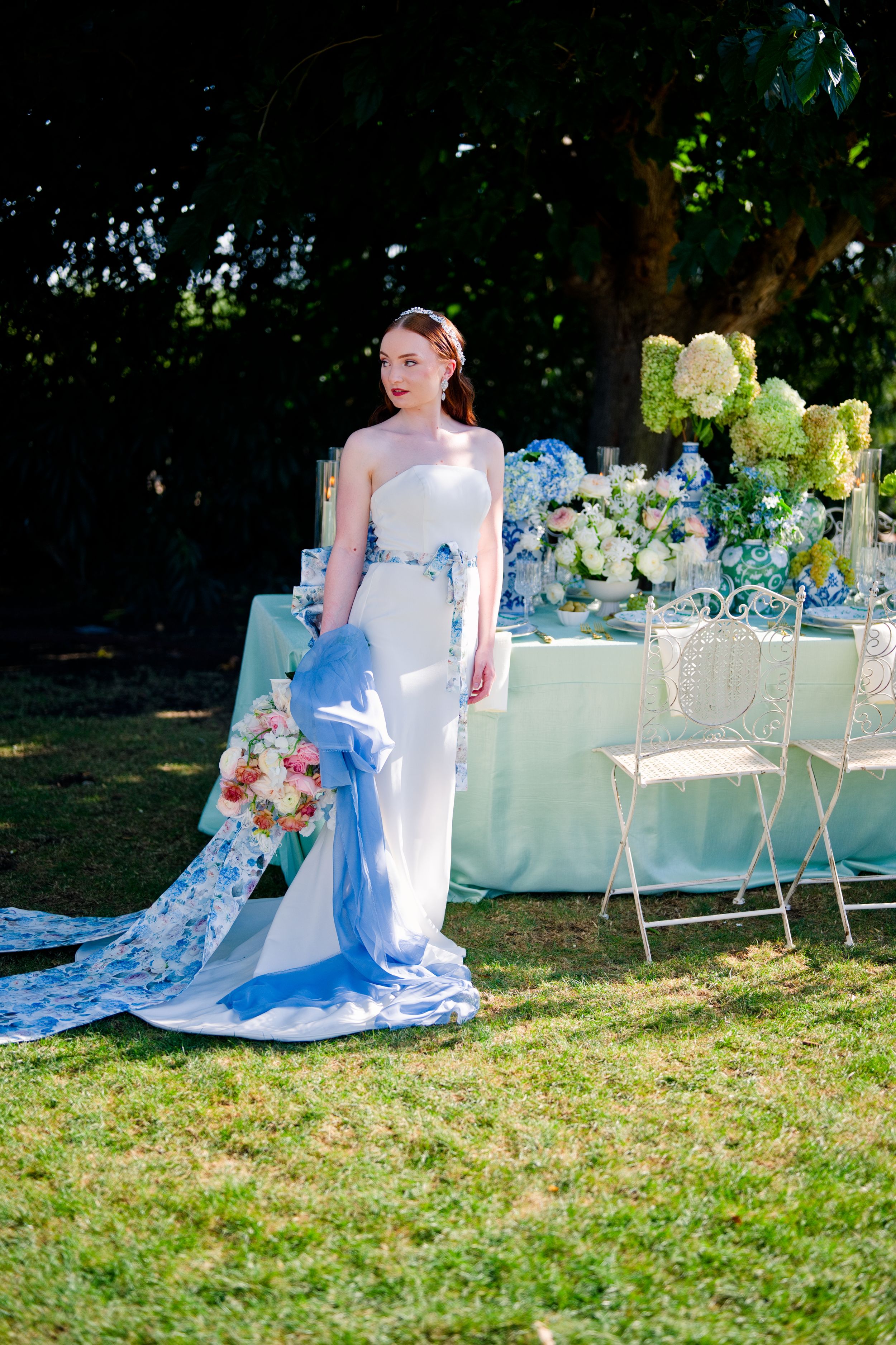 Elegant bride in white gown with blue accents poses by garden reception table with floral arrangements.