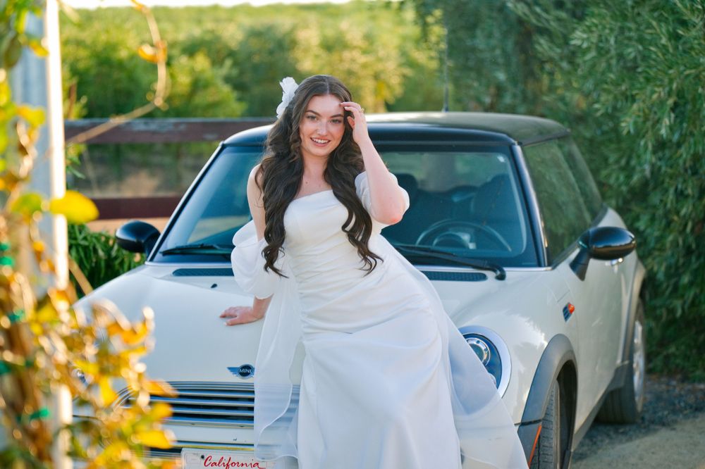 Bride in flowing white wedding dress posing on a Mini Cooper on a sunny day.