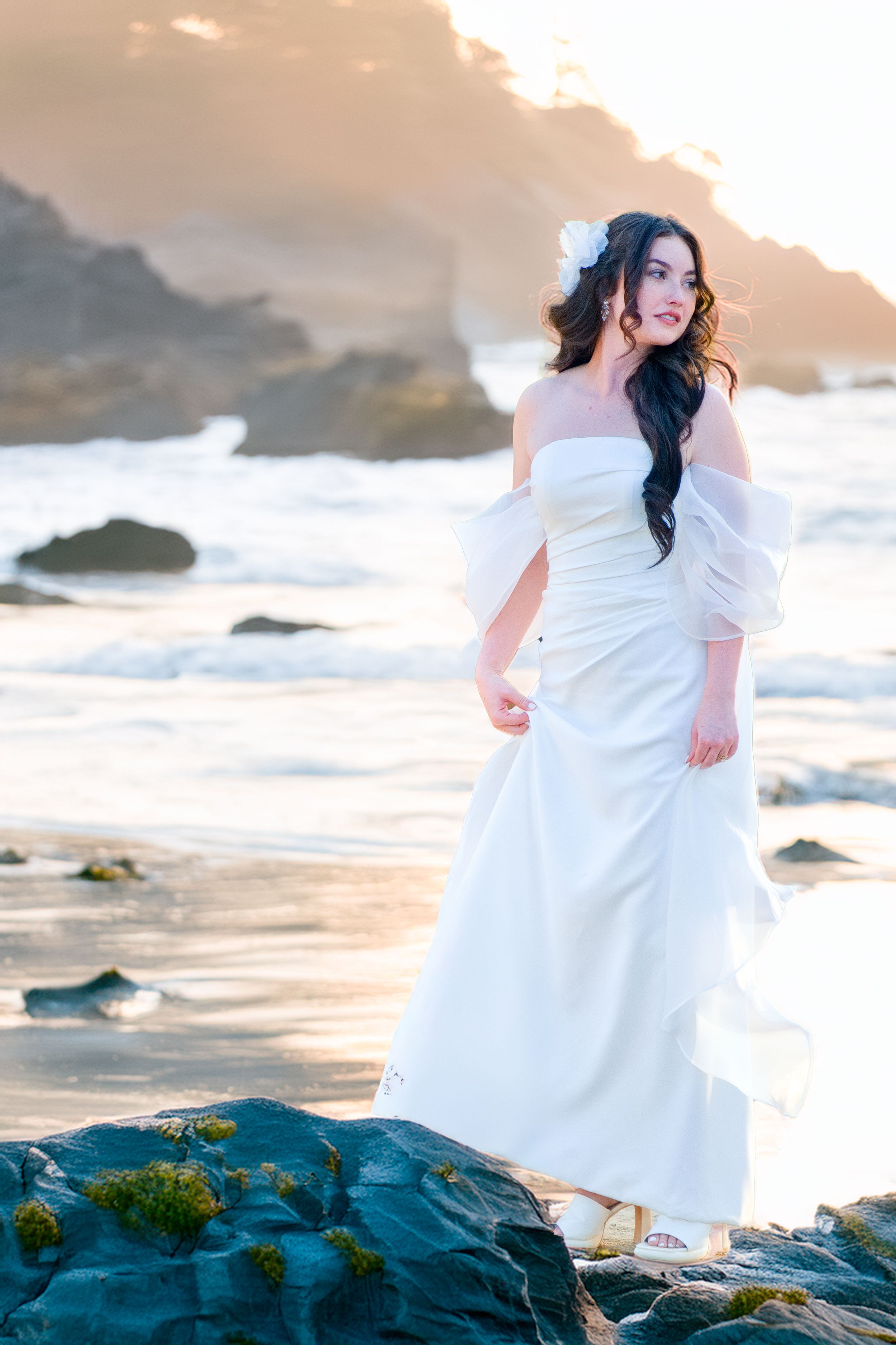 A bride in a flowing white off-shoulder bridal gown stands on coastal rocks at sunset with waves crashing in the background.
