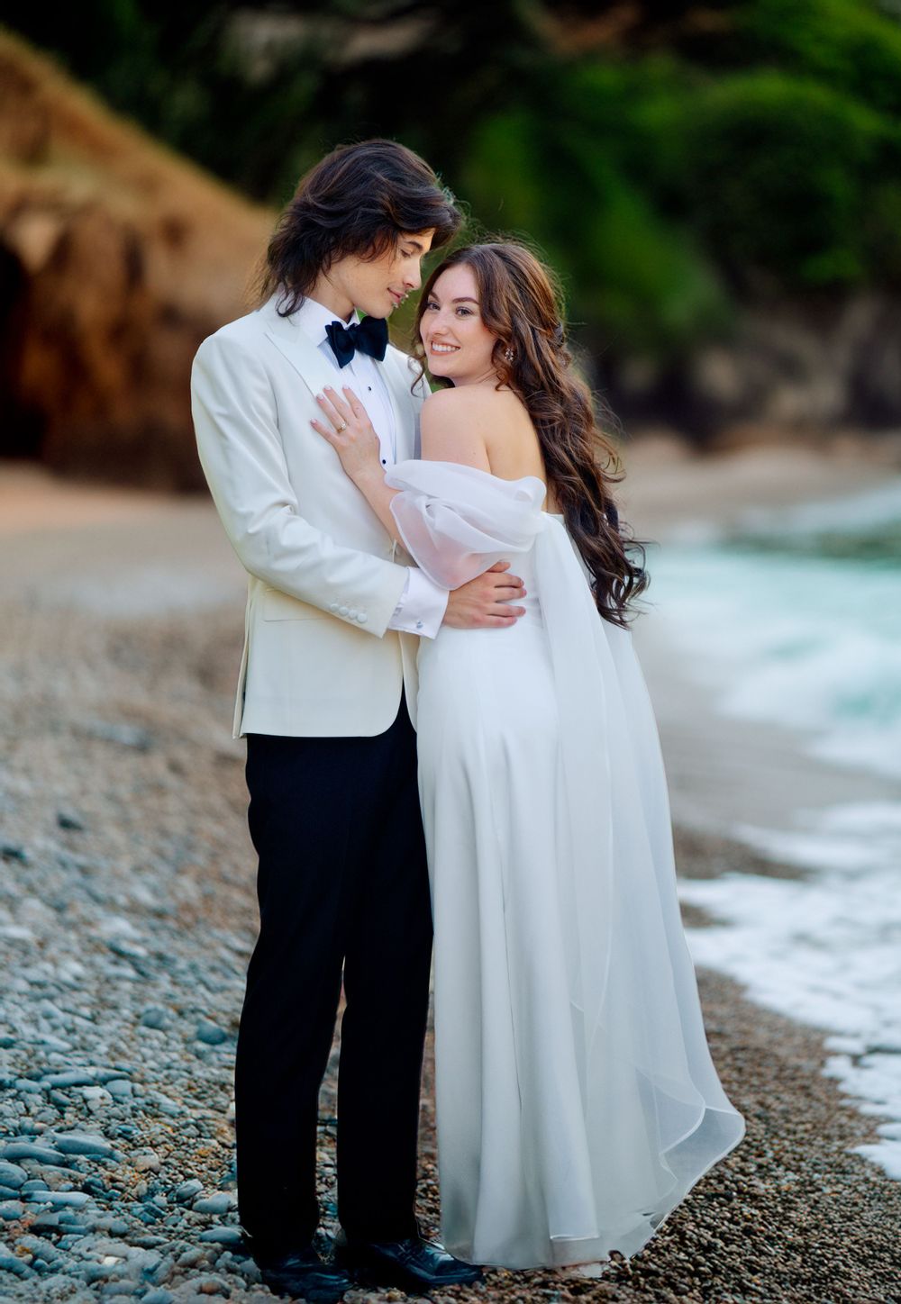 Couple in formal wedding attire embracing on rocky beach.