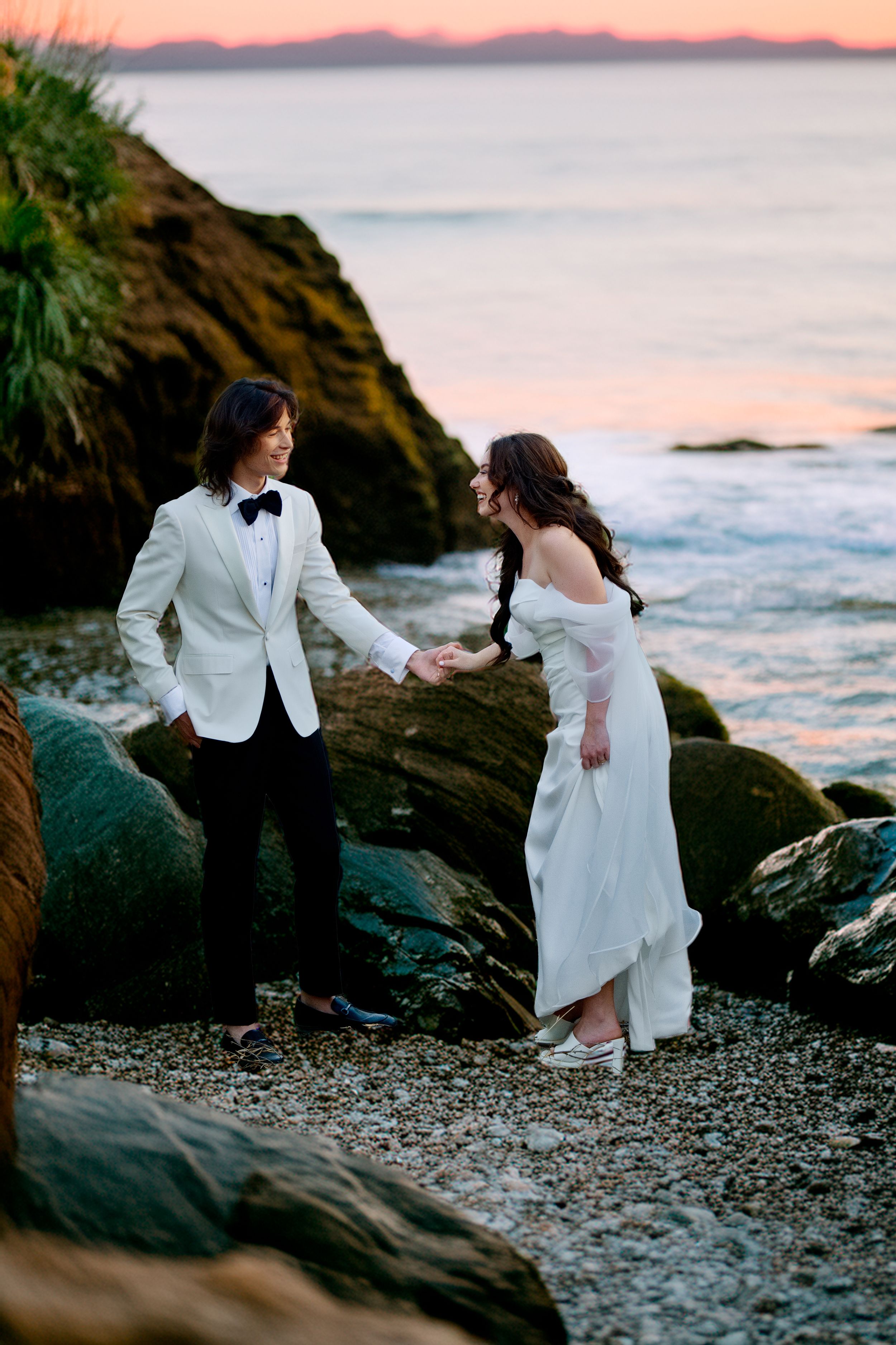 A newlywed couple in wedding attire holds hands on a rocky beach at sunset with waves and pink sky in the background.