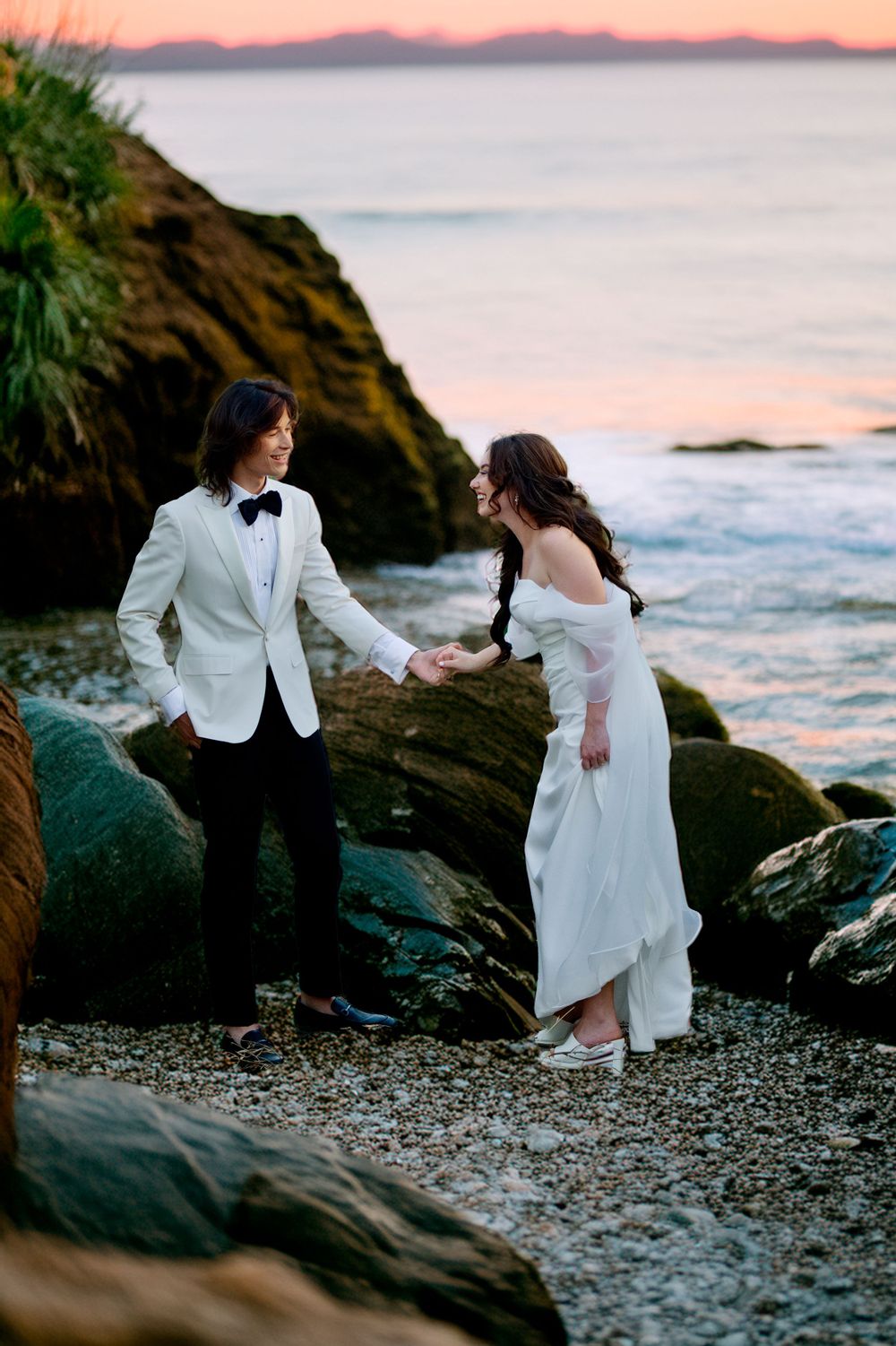 A newlywed couple in wedding attire holds hands on a rocky beach at sunset with waves and pink sky in the background.