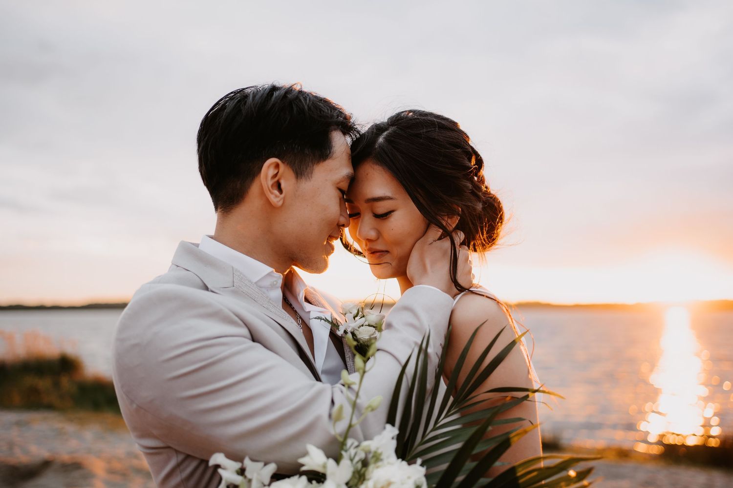 A couple shares an intimate moment at sunset by the ocean with palm leaves and flowers in their romantic photoshoot.