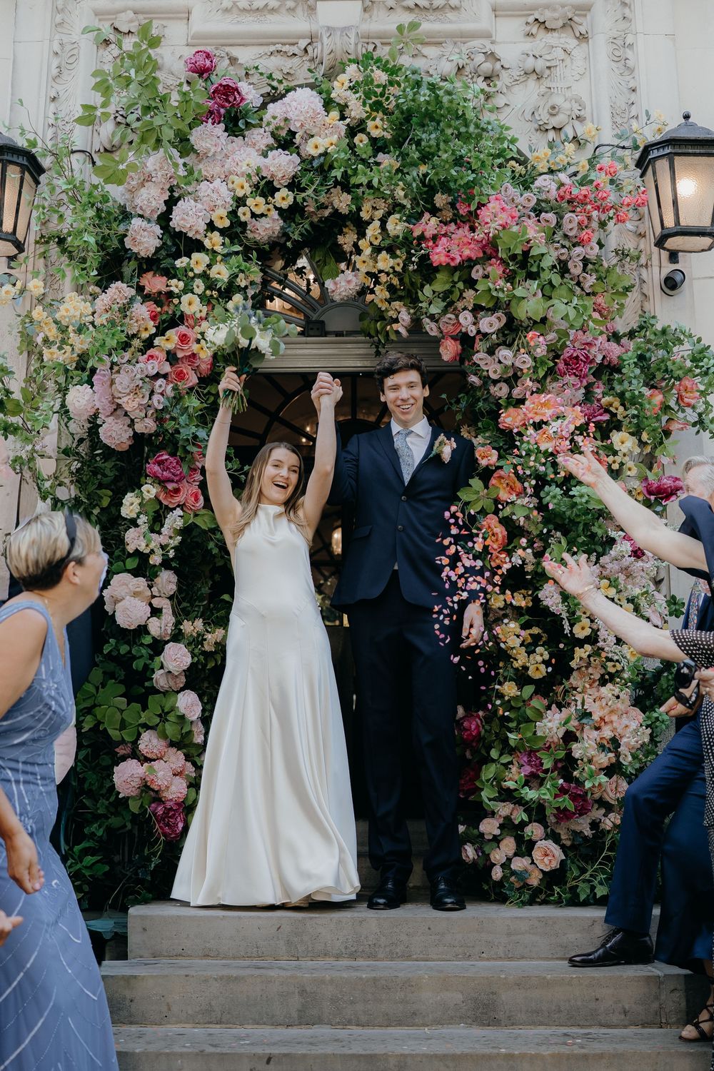 Joyful bride and groom raise clasped hands beneath a lush pink and orange floral arch as wedding guests cheer on the steps.
