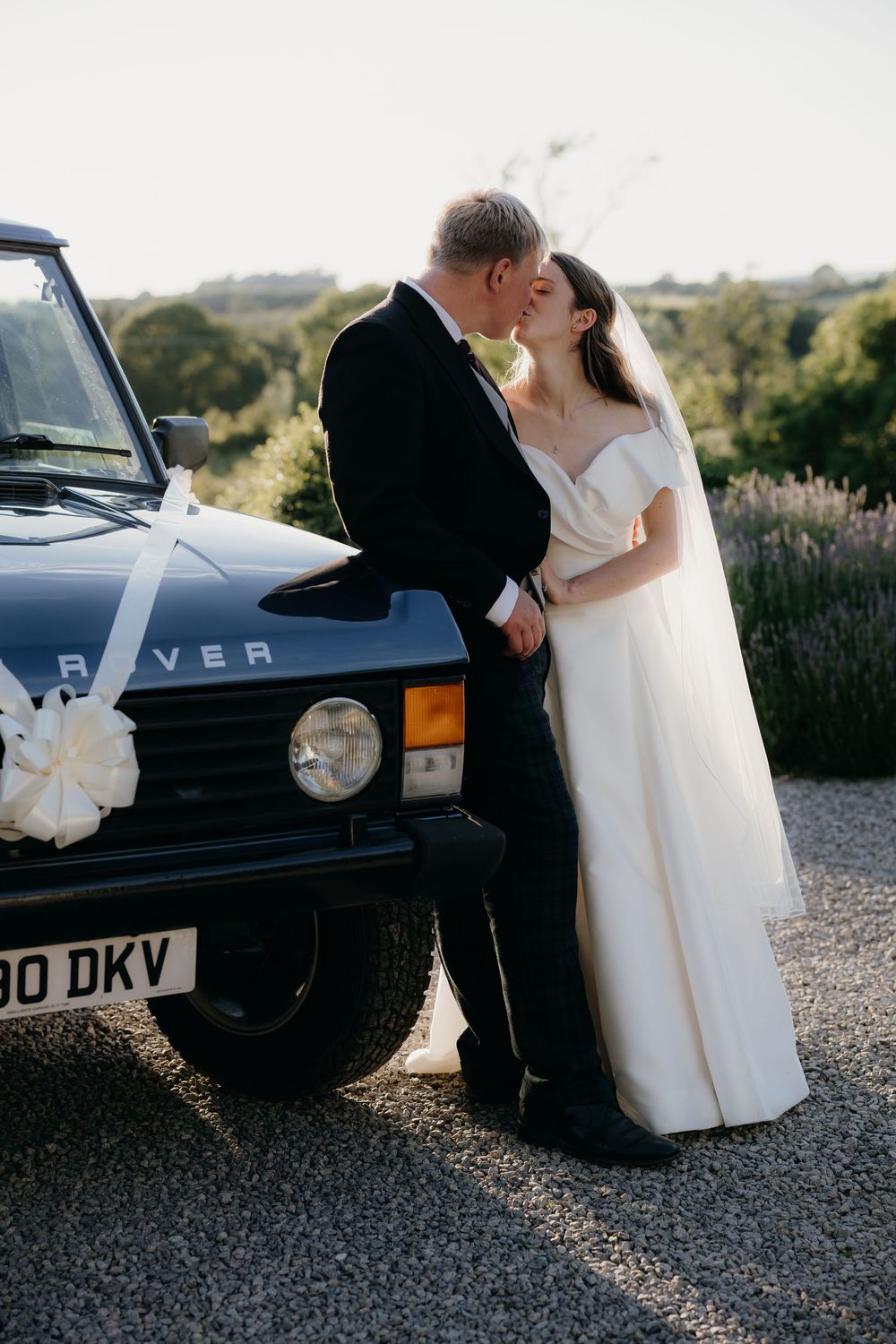Bride in an off-shoulder white gown and veil kisses the groom beside a vintage Range Rover decorated with a white ribbon bow.