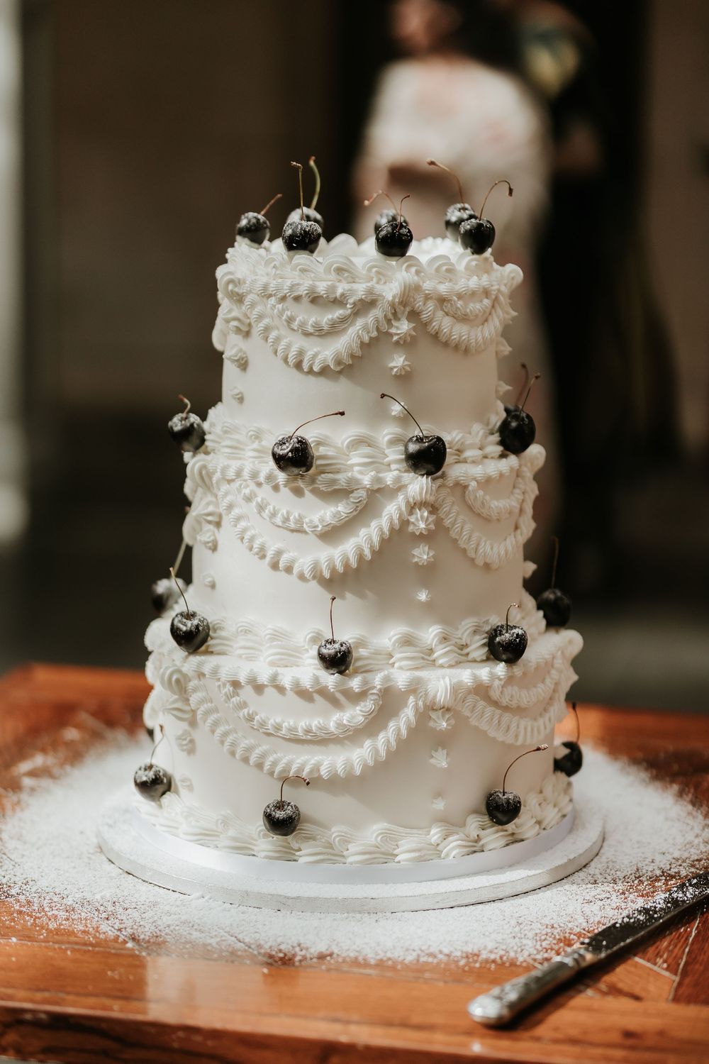 Three-tiered white wedding cake decorated with delicate lace piping and fresh black cherries on a rustic wooden surface.