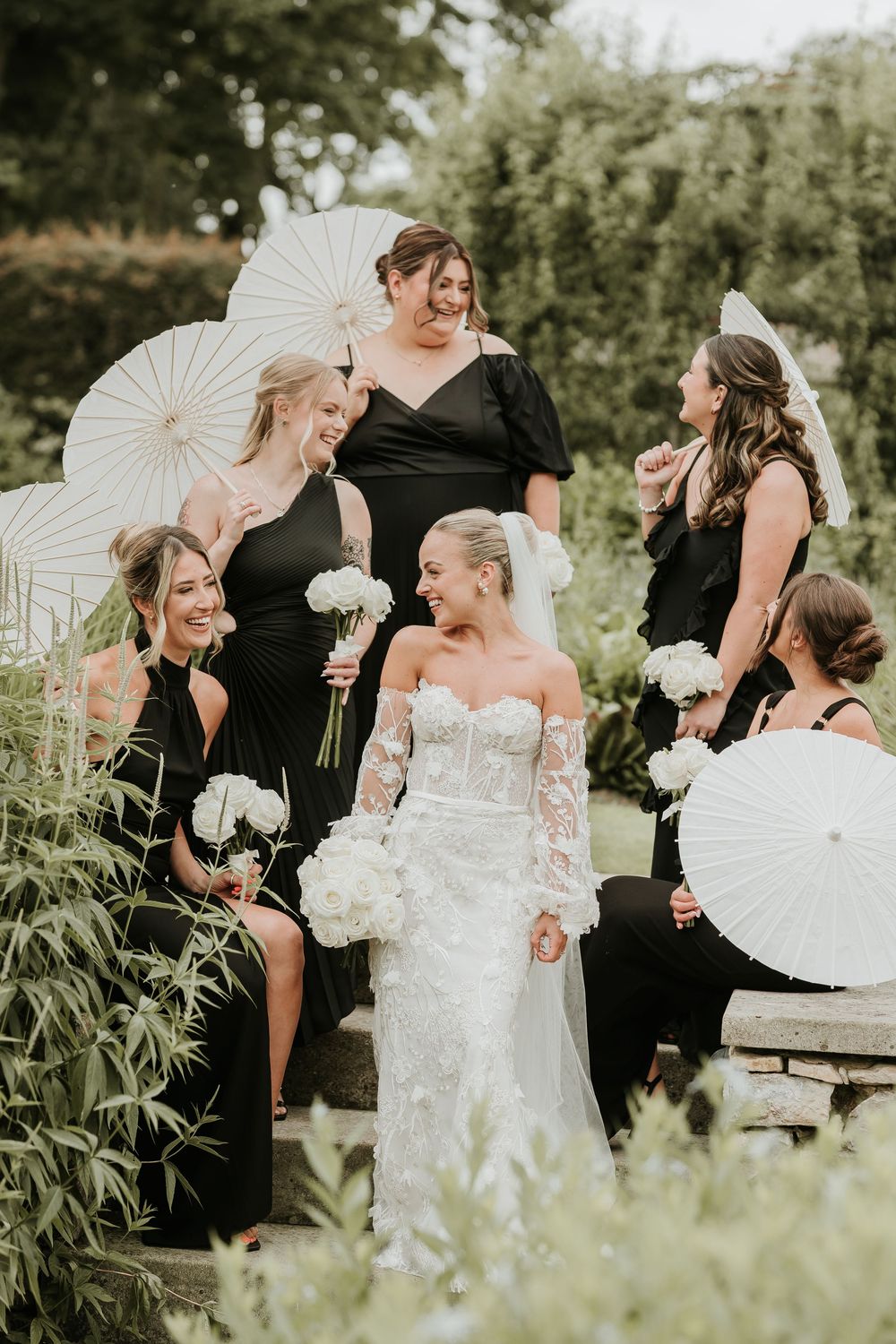 Bride in lace gown with bridesmaids in black dresses holding white parasols and floral bouquets outdoors.