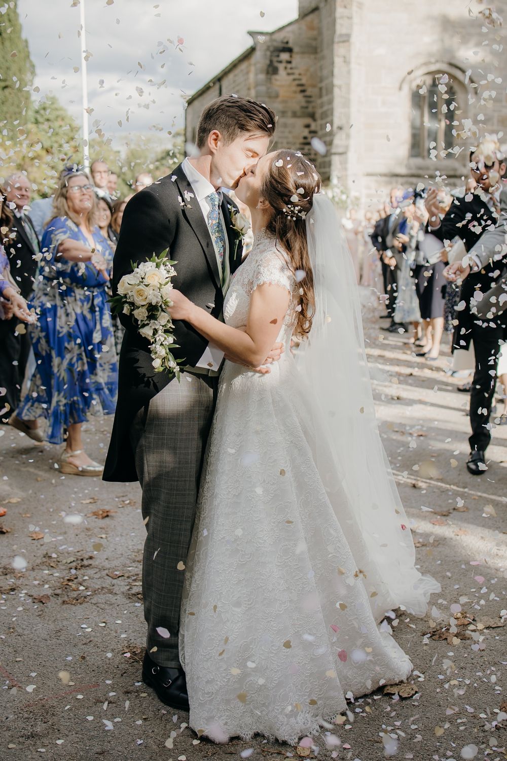Bride and groom share a kiss while colorful confetti falls around them outside a stone church.