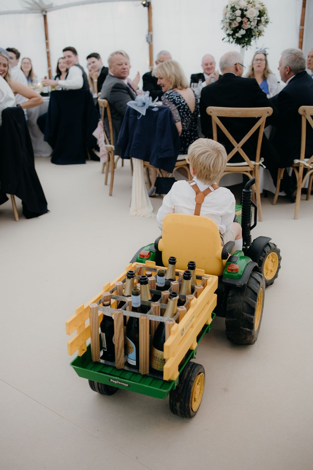 Young boy driving a John Deere toy tractor delivering bottles at a wedding reception in a white tent.