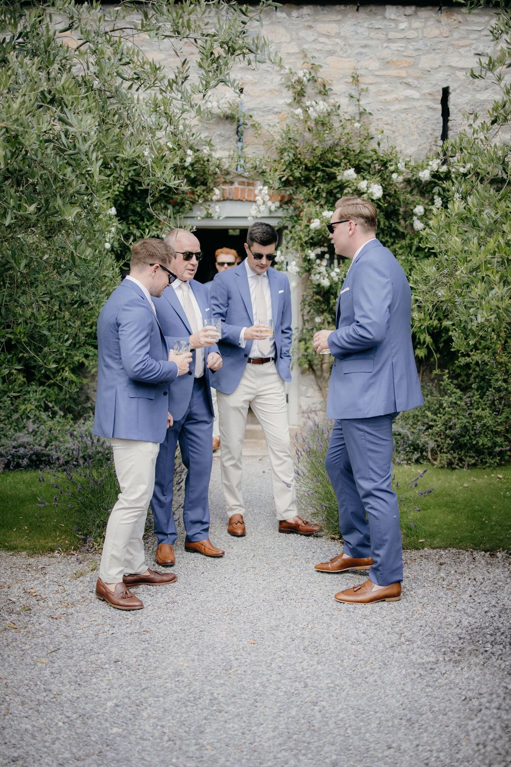 Four groomsmen in blue suits and tan chinos socializing on a gravel path at an elegant garden wedding venue.
