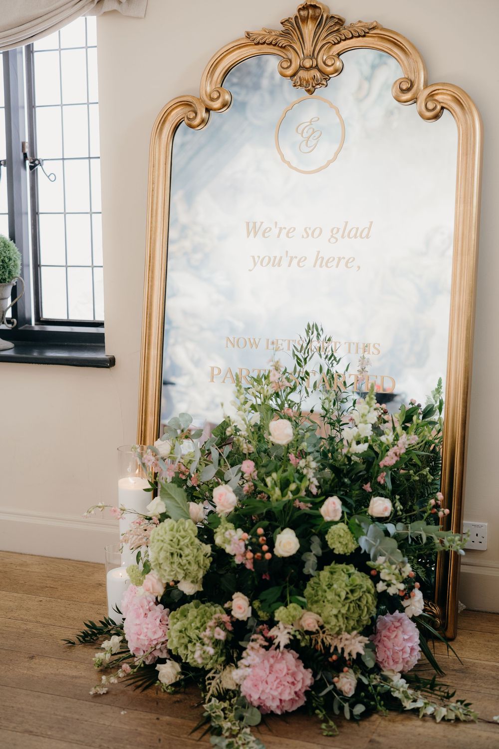 Ornate gold-framed wedding welcome mirror reading "We're so glad you're here" beside a lush pink and green hydrangea floral arrangement.