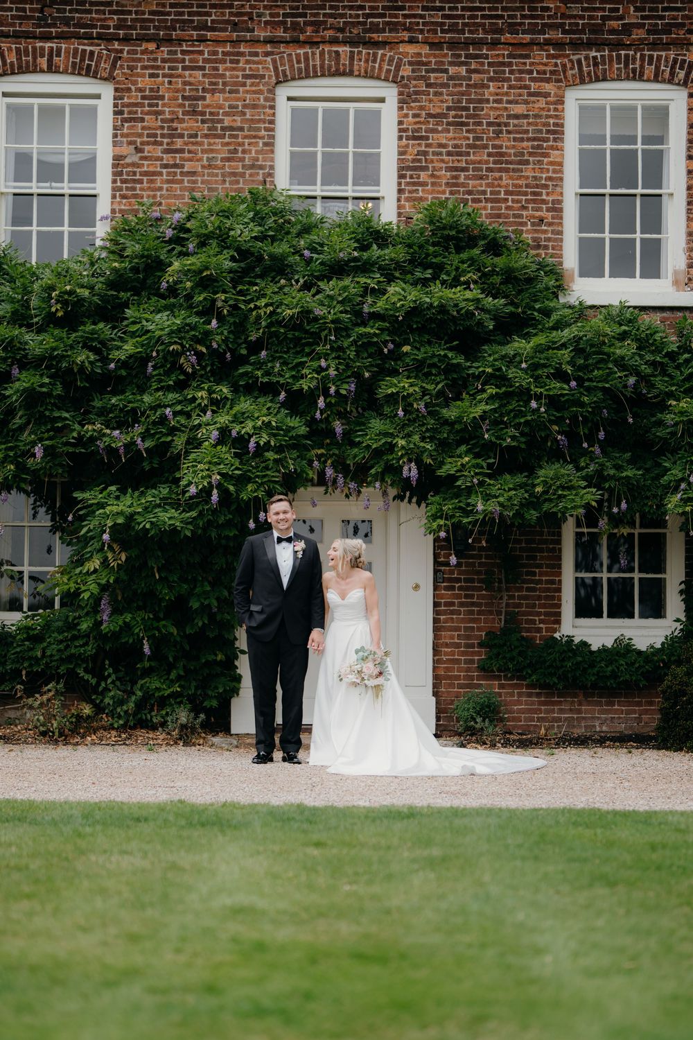 Bride in a white gown and groom in a black tuxedo hold hands before a red brick manor house covered in wisteria on a sunny lawn.