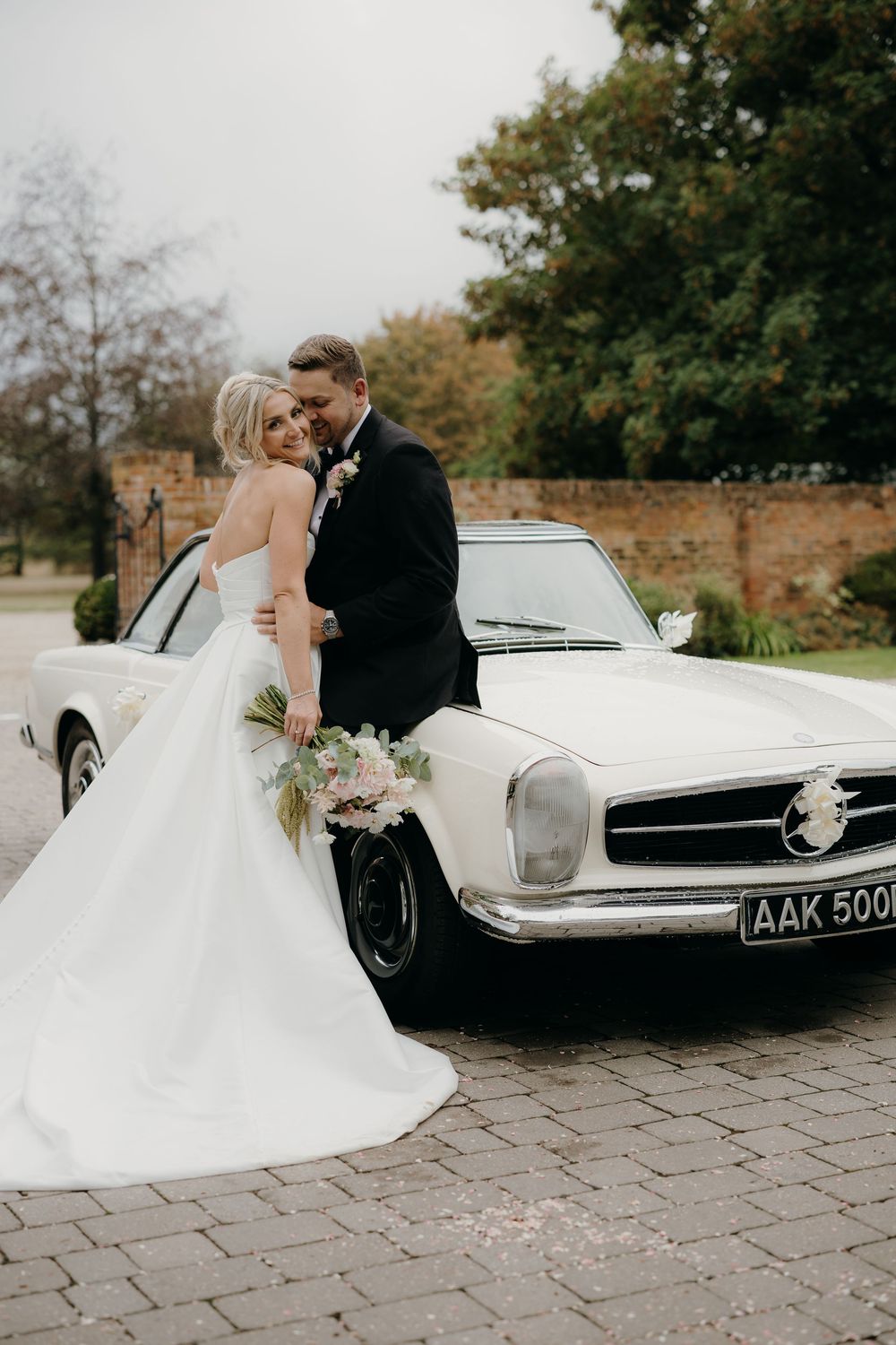 Bride in a white gown and groom in a black suit embrace beside a vintage white Mercedes convertible on a cobblestone driveway.