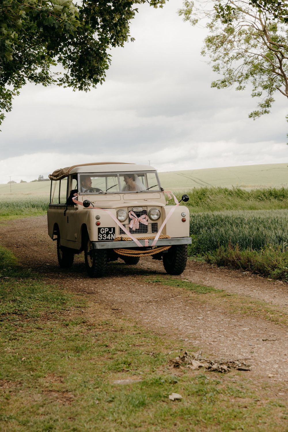 Vintage cream Land Rover decorated with wedding ribbons driving along a rural dirt path surrounded by green fields.