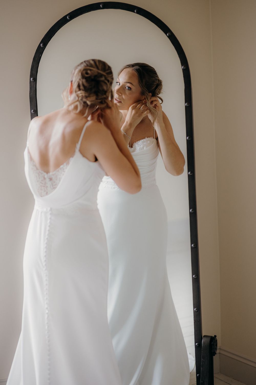 Bride in a white wedding gown with lace back detail adjusts her earring while admiring her reflection in a tall arched black-framed mirror.