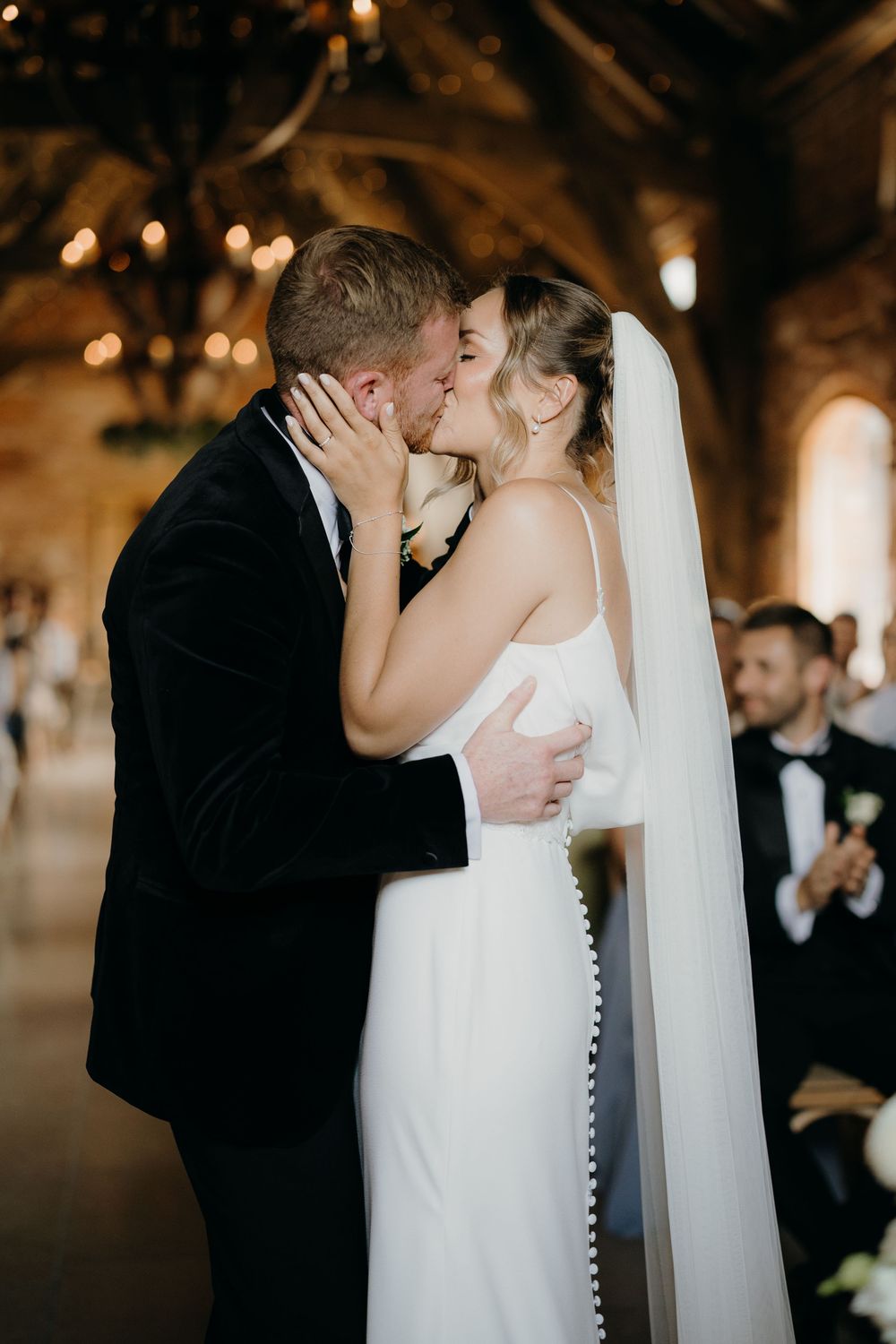 Bride in a white gown and veil kisses groom in a black velvet jacket during a wedding ceremony in a rustic venue with chandeliers.