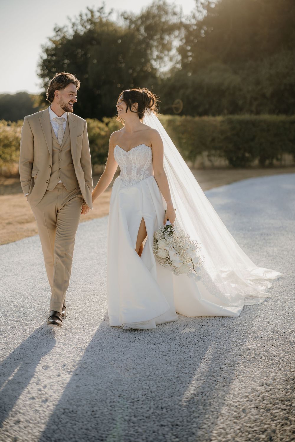 Bride in a strapless white gown with long veil holding a bouquet walks hand in hand with a smiling groom in a tan suit on a gravel path.