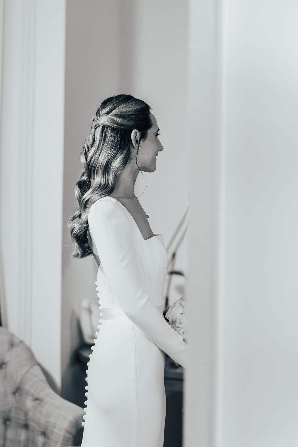 Black and white side profile portrait showing elegant wedding dress with long sleeves and flowing wavy hairstyle.