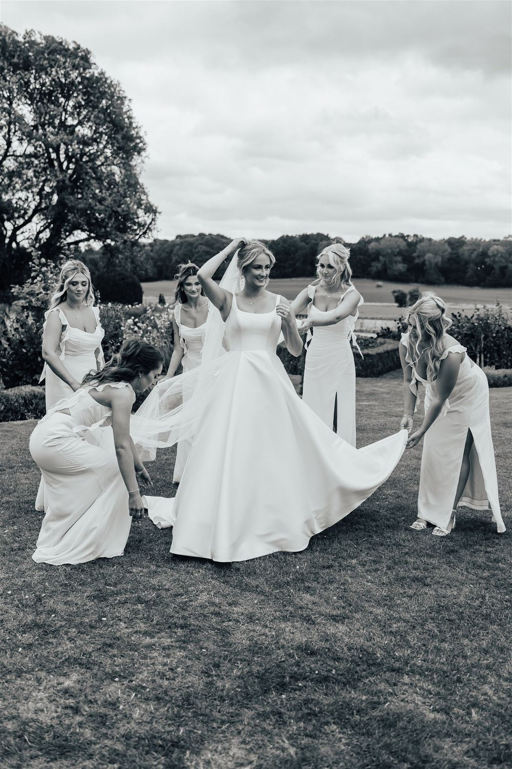 A group in white dresses poses playfully in sequence across a grassy field under cloudy skies.