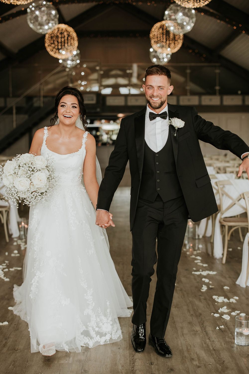 Elegant wedding couple walking together in a rustic venue decorated with string lights and white floral arrangements.