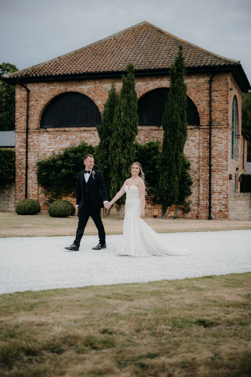 Bride in a white gown and groom in a black tuxedo hold hands walking along a gravel path past a rustic brick building and cypress trees.