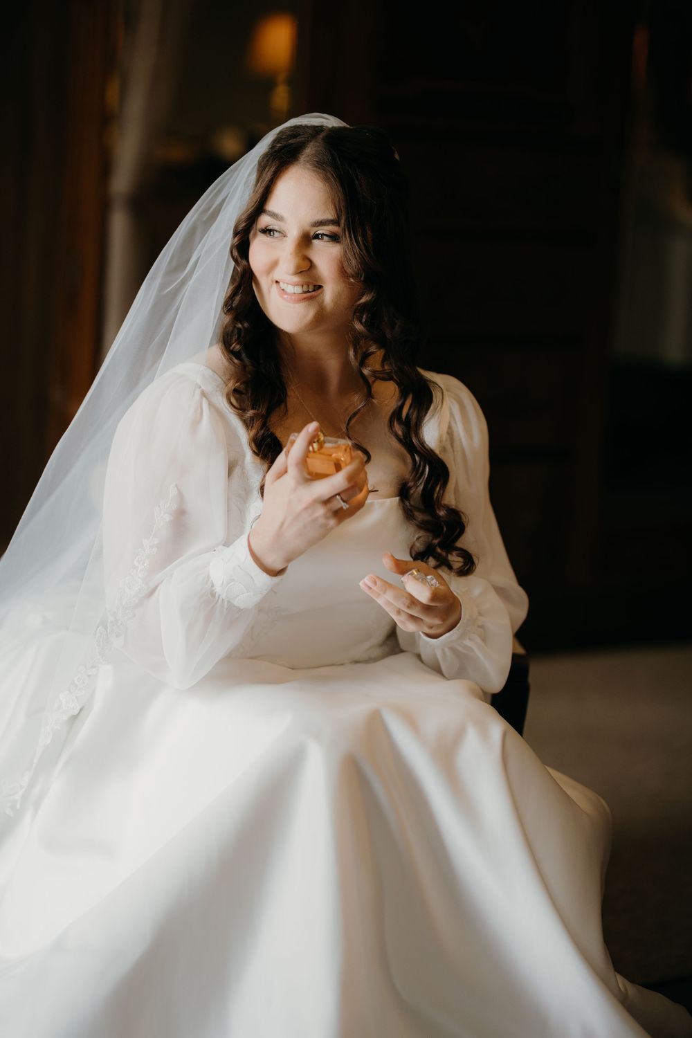 Smiling bride in an elegant long-sleeved white wedding gown and veil sits holding a drink in a softly lit room.