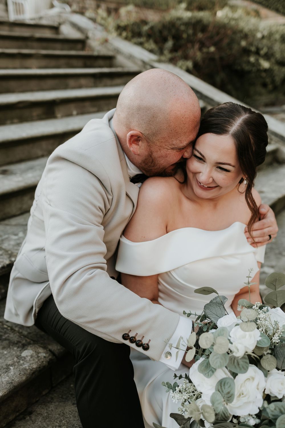 A wedding couple shares an intimate moment on stone steps in a garden setting, surrounded by white floral bouquets.