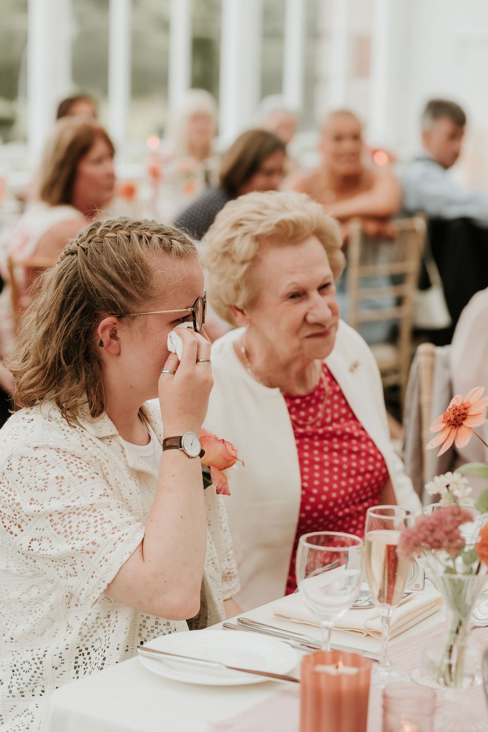 Two people share a moment at an elegant dining table with floral centerpieces and glowing candlelight during a celebration.