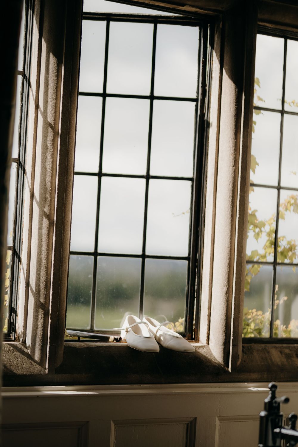 White flat bridal shoes rest on a stone windowsill beside an open leaded window with sunlit green countryside beyond.
