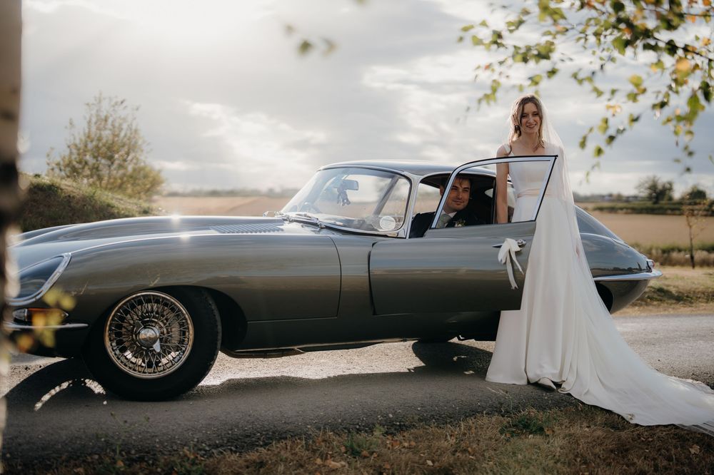 Bride in a flowing white wedding gown standing beside a vintage gray Jaguar E-Type convertible on a country road at sunset.