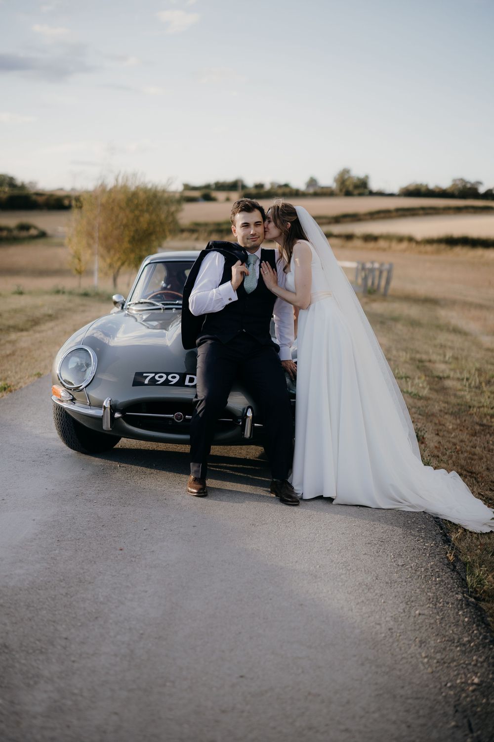 Bride in a flowing white gown and groom in a vest lean on a vintage grey sports car on a country road at sunset.