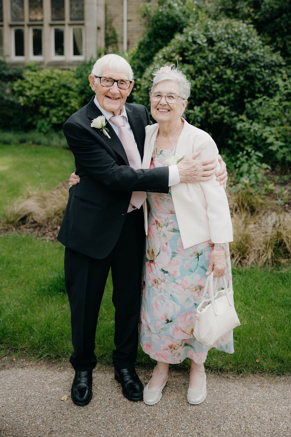 Elderly couple embracing outdoors at a wedding, man in dark suit with boutonniere, woman in floral dress and white jacket.